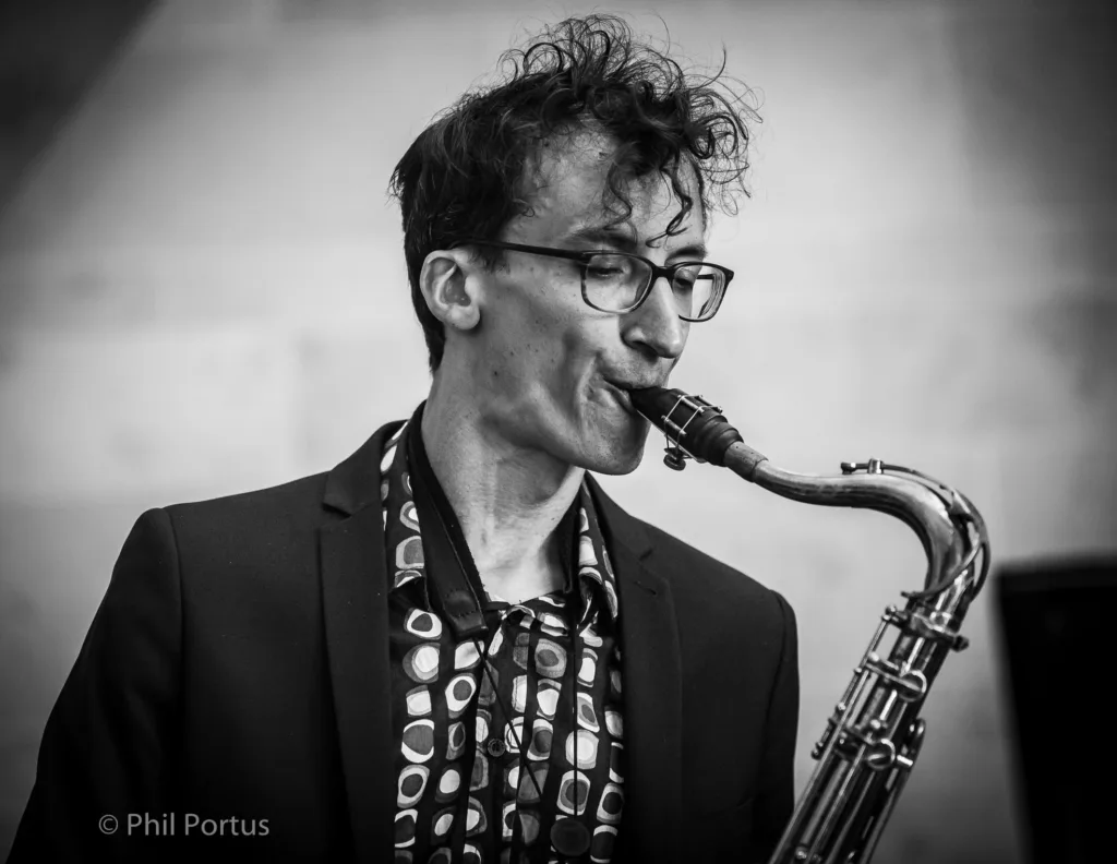 A musician plays a saxophone while wearing glasses and a patterned shirt under a suit jacket.