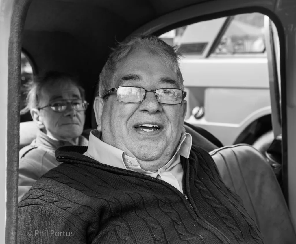 Two men sit inside a car; the man in front smiles and wears glasses.