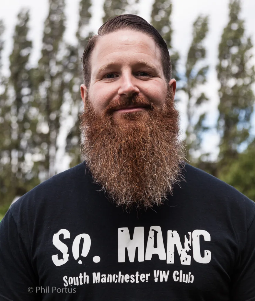 A man with a full beard smiles directly at the camera while wearing a black t-shirt.