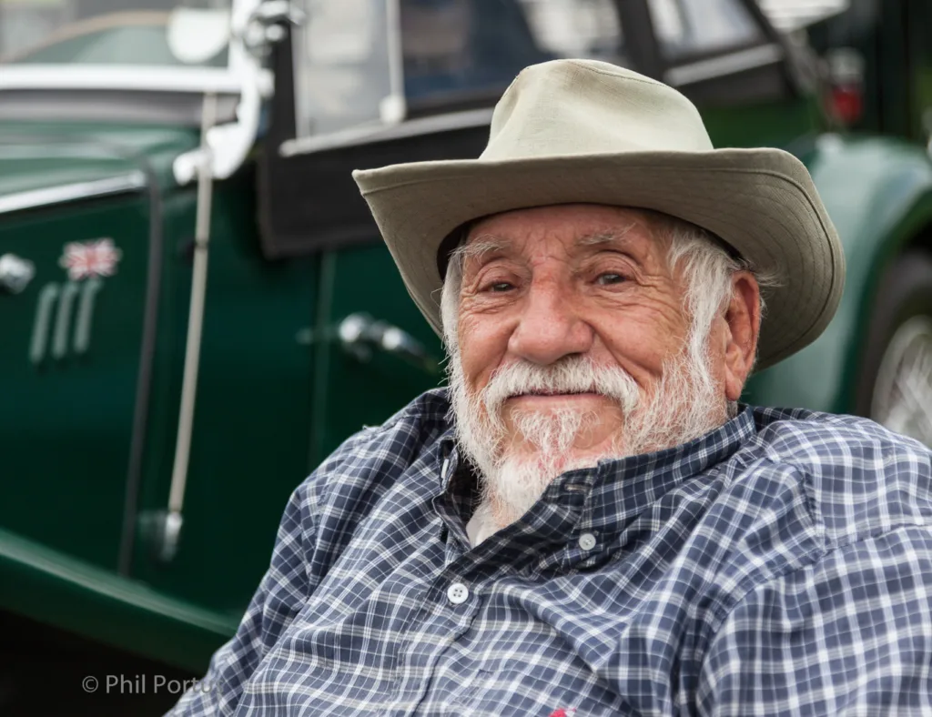 An older man with a full white beard wears a light brown hat and plaid shirt, leaning against a dark green vehicle.