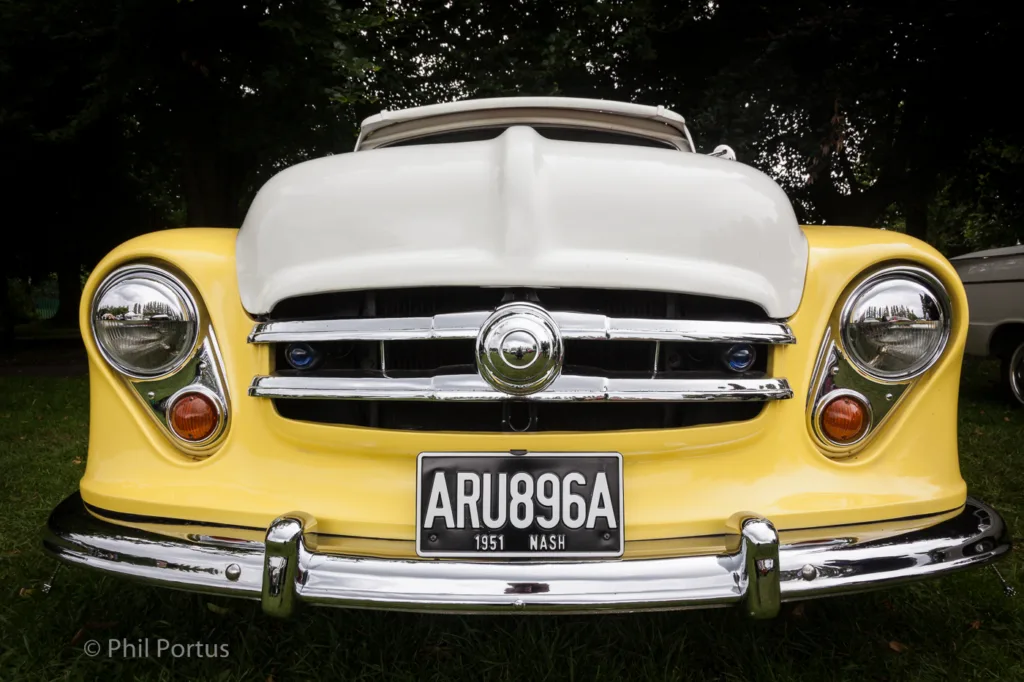 A yellow car with a white hood and chrome accents sits on green grass under trees.