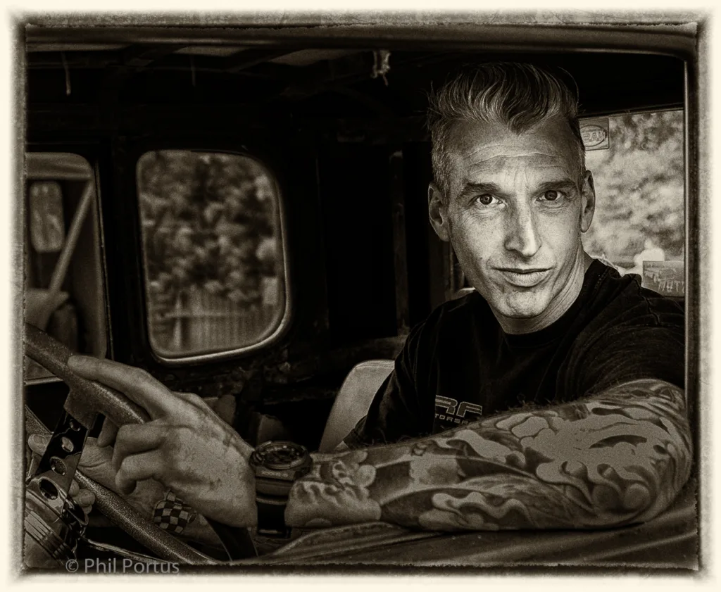 A man with a stylized haircut looks directly at the camera while seated in a vintage vehicle.