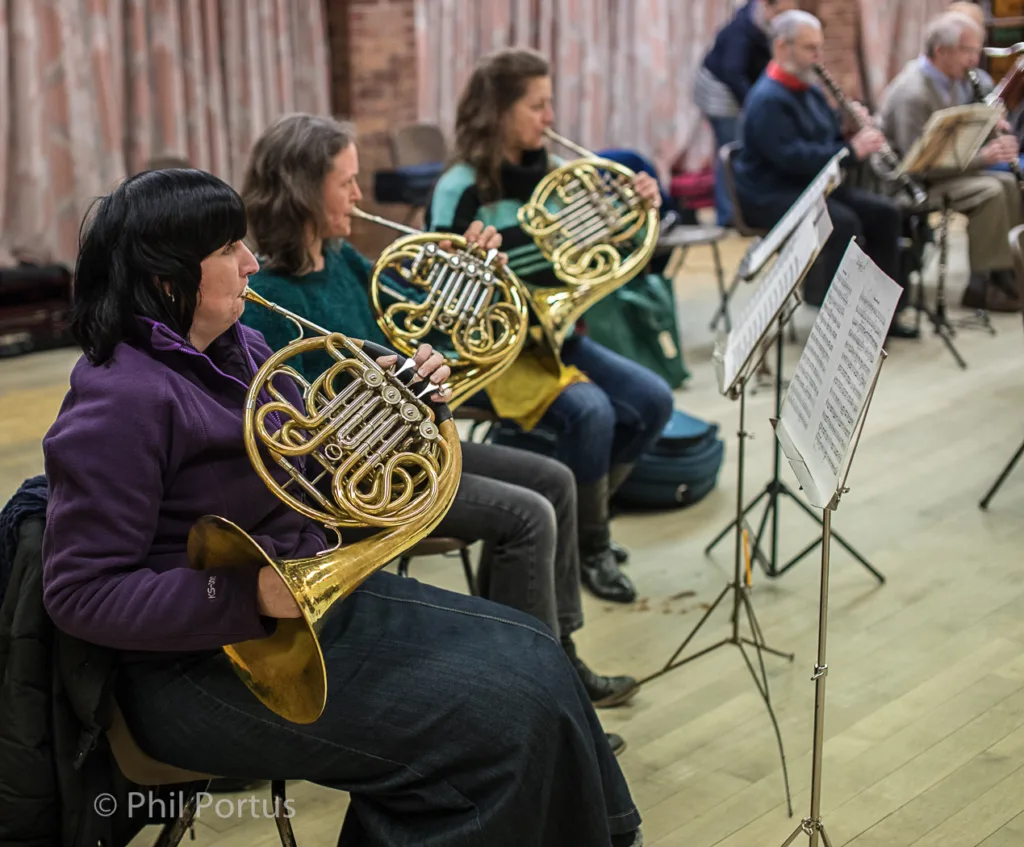 Several people play brass instruments during a rehearsal in a room with wooden floors and dark curtains.