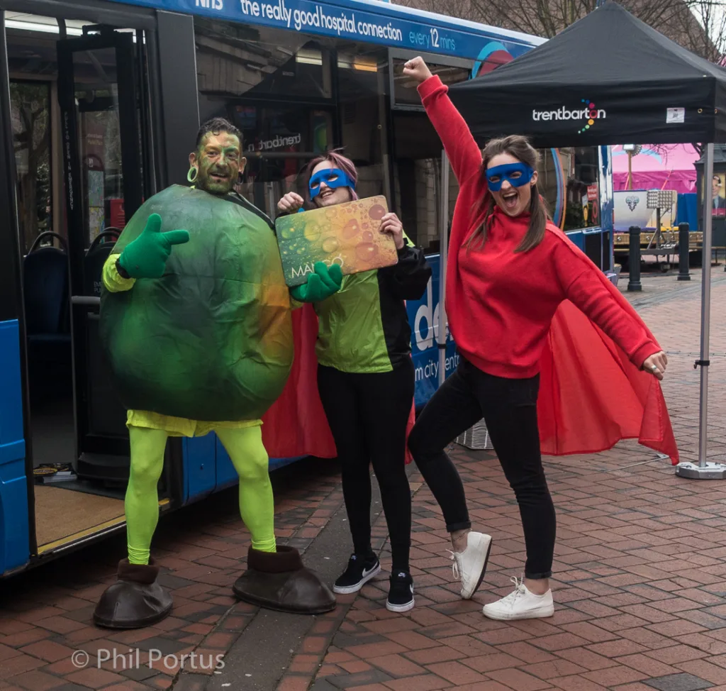 Three people pose outdoors; two wear superhero costumes with blue masks and capes, one wears a large green turtle costume.