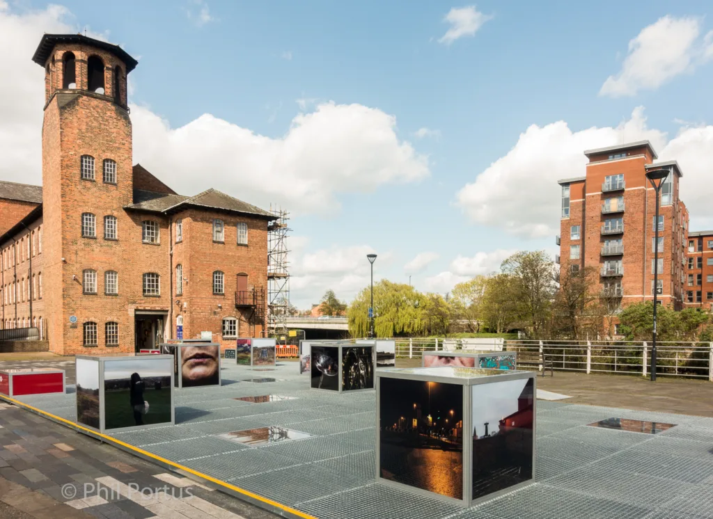 Several metal cubes displaying images are arranged on a paved area in front of brick buildings and modern apartments.