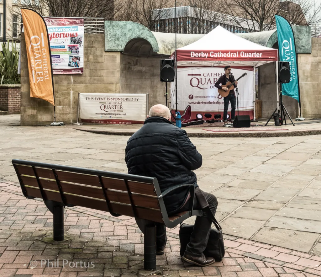 A person sits on a bench facing a musician playing guitar under a red and blue awning.