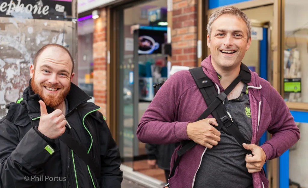 Two smiling men pose for a portrait outdoors, one giving a thumbs up.