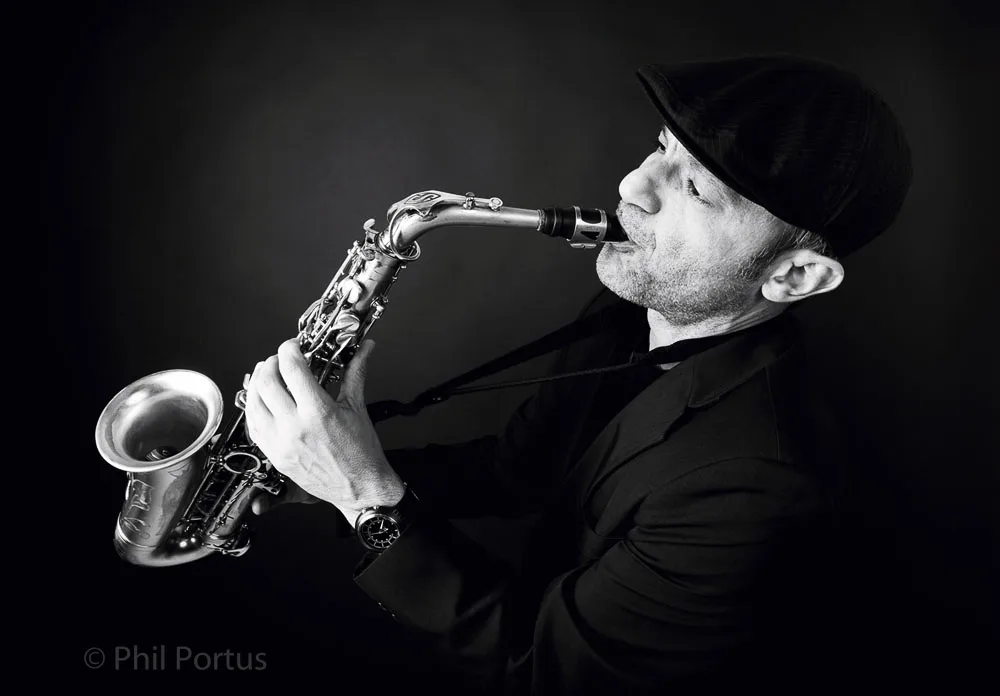 A person wearing a hat and dark clothing plays a saxophone in black and white.