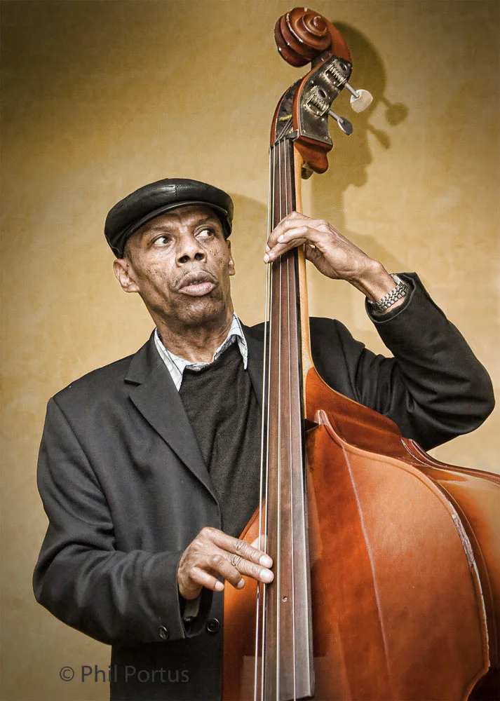 A musician stands holding an upright bass, wearing a suit jacket and cap.