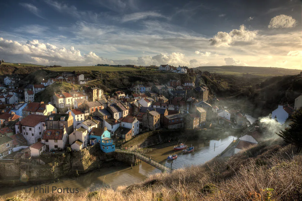 A hillside village with colorful buildings sits nestled in a valley beneath a cloudy sky.