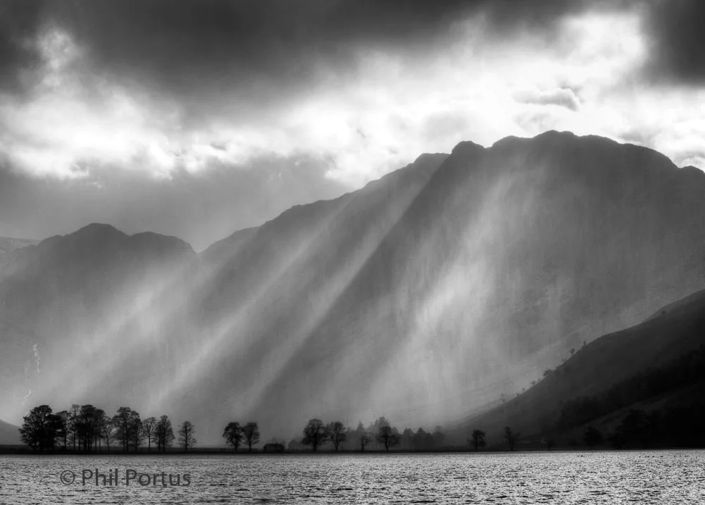 Black and white landscape shows mountains rising from a body of water with dramatic rays of light.