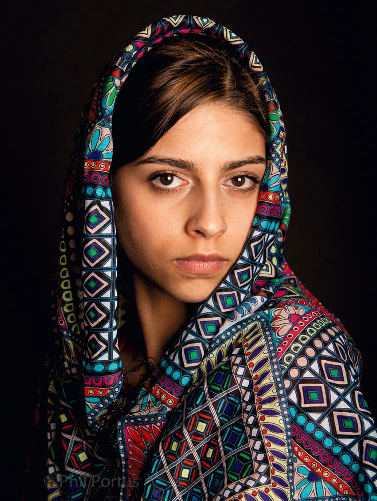 A young person with dark hair looks directly at the viewer, draped in a colorful patterned scarf.