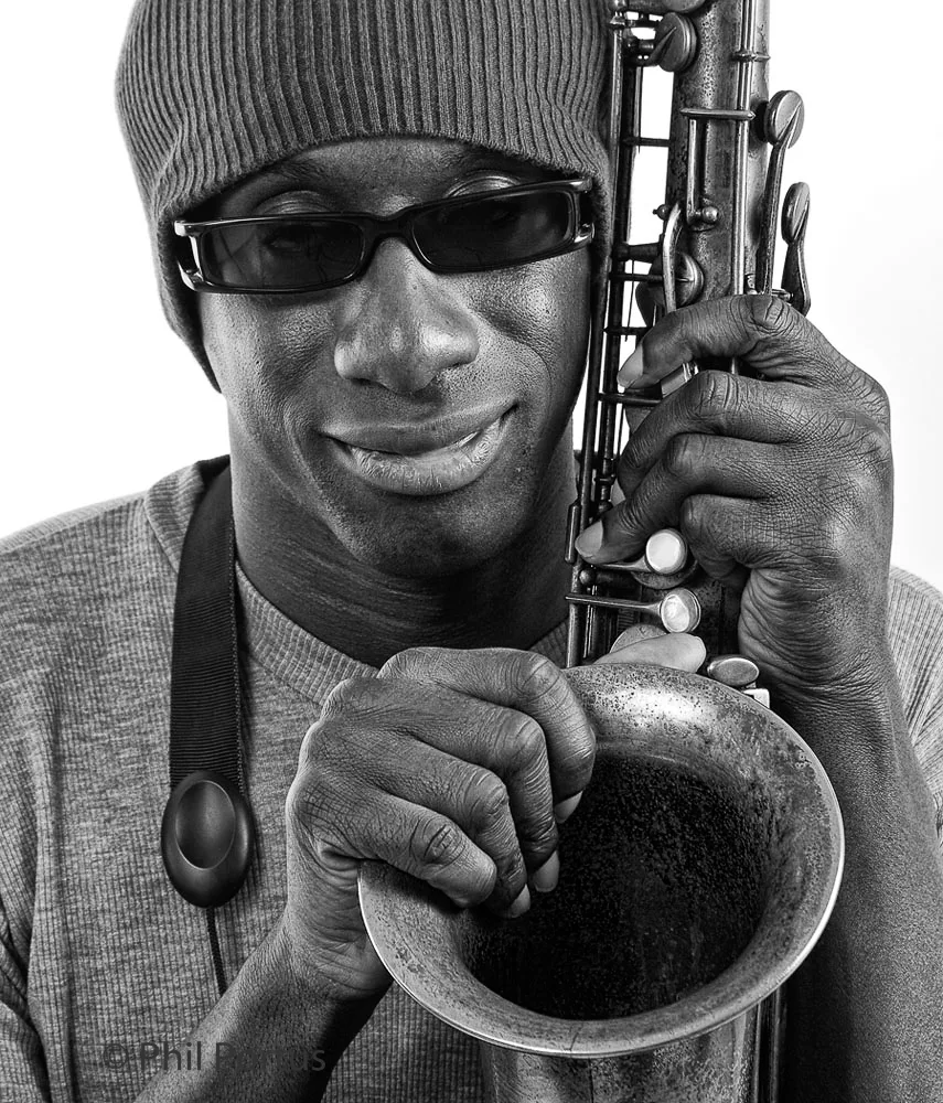A man wearing sunglasses and a knit cap holds a saxophone with his hands.