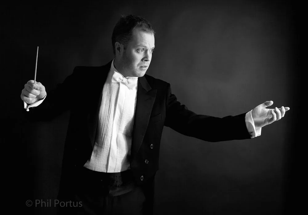 A man in formal wear holds a baton while gesturing with his hand against a dark background.