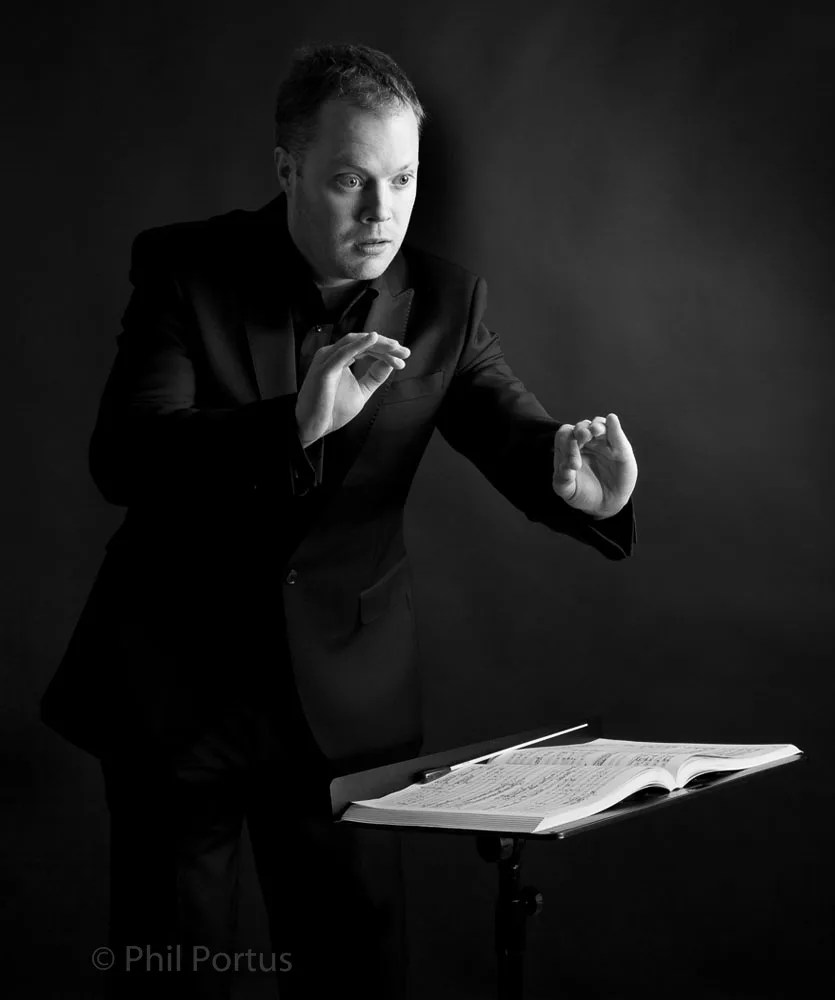 A person in a dark suit gestures with their hands while standing next to a music stand.