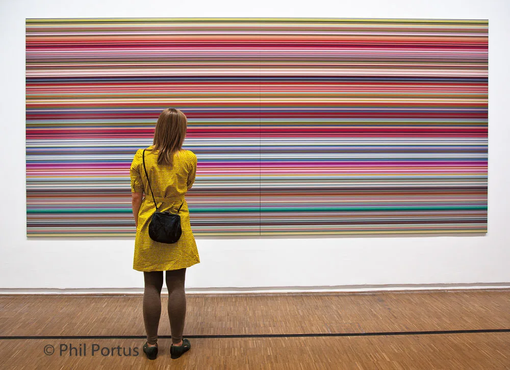 A person with red hair stands facing a large rectangular artwork composed of horizontal colored stripes.