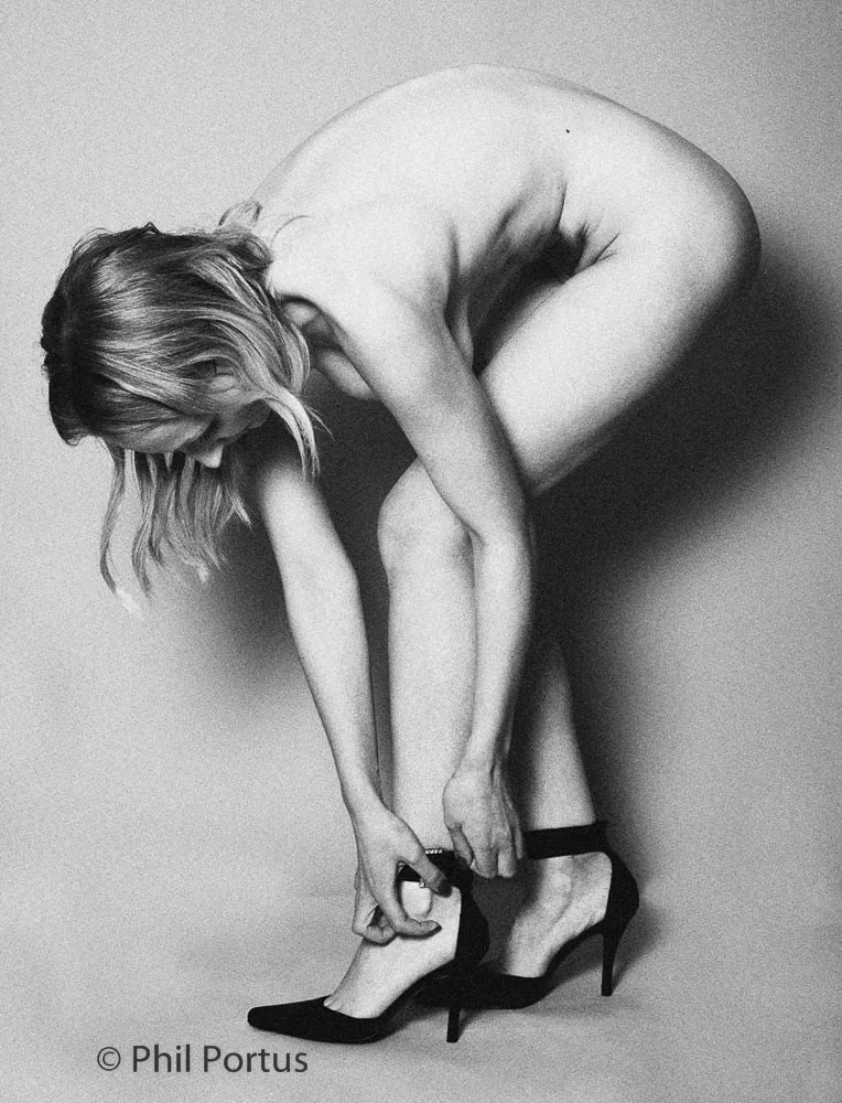 A person bends forward, reaching for a pair of black high heels while appearing unclothed.