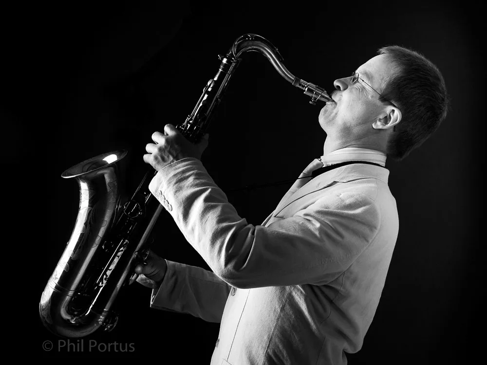 A person wearing glasses plays a saxophone while dressed in a light-colored suit jacket.