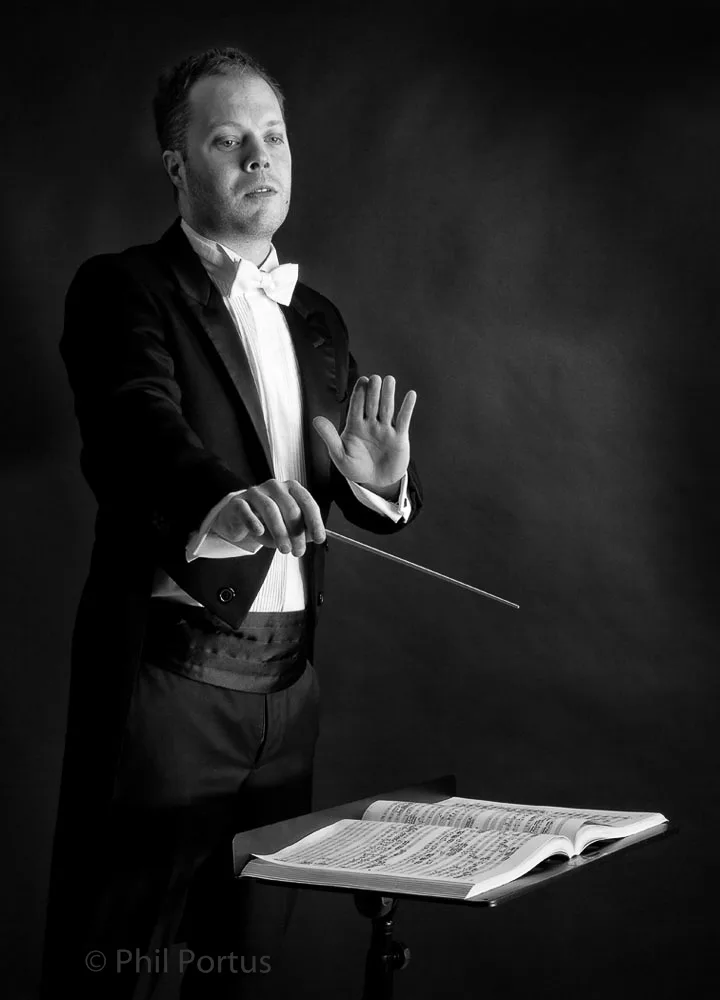 A man in a formal black suit and bow tie conducts with a baton, sheet music visible on a stand.