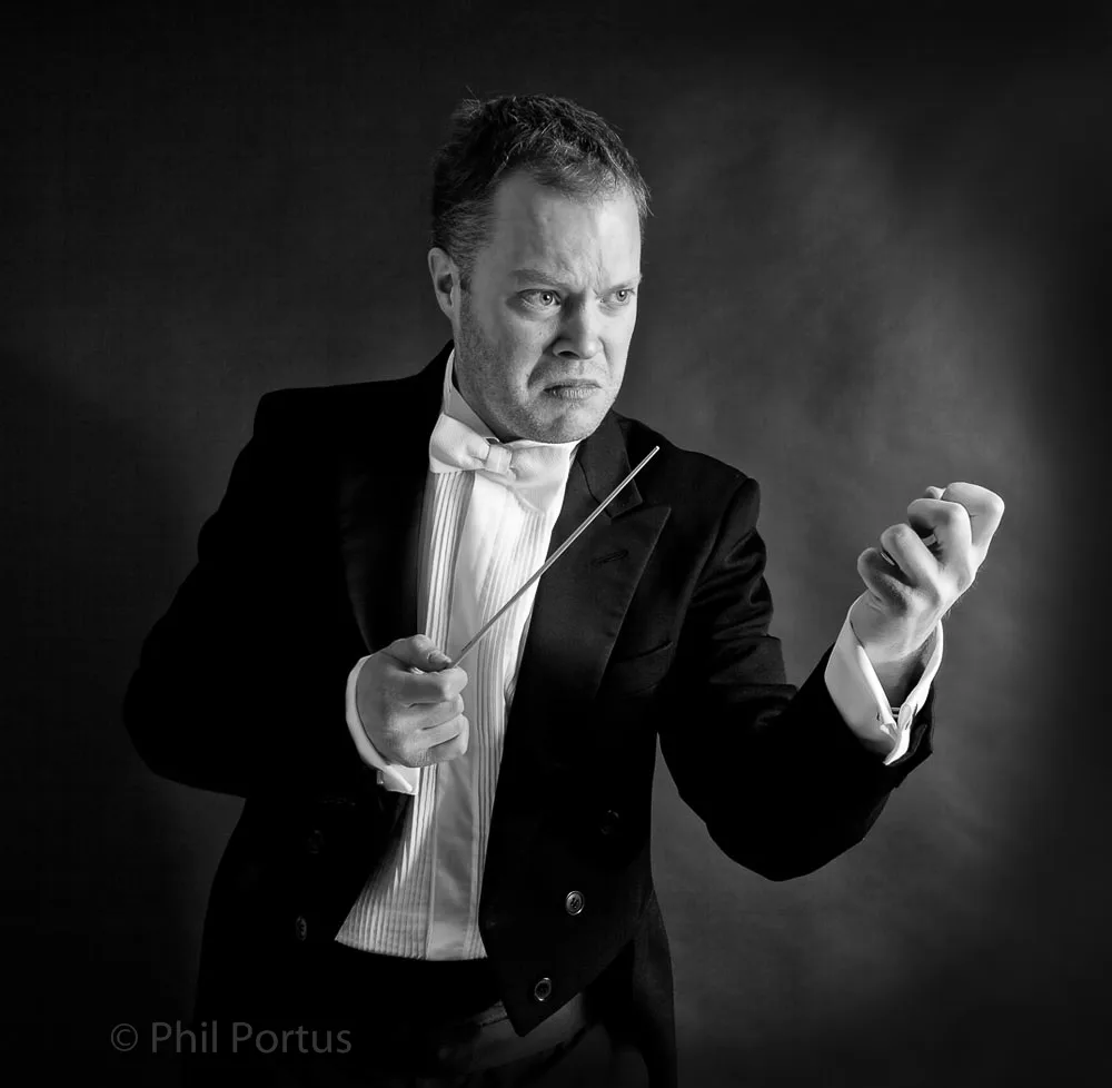 A man in a tuxedo holds a baton while gesturing with his hands.