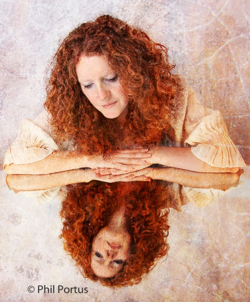 A woman with curly red hair rests her arms on a reflective surface, creating a mirrored image.