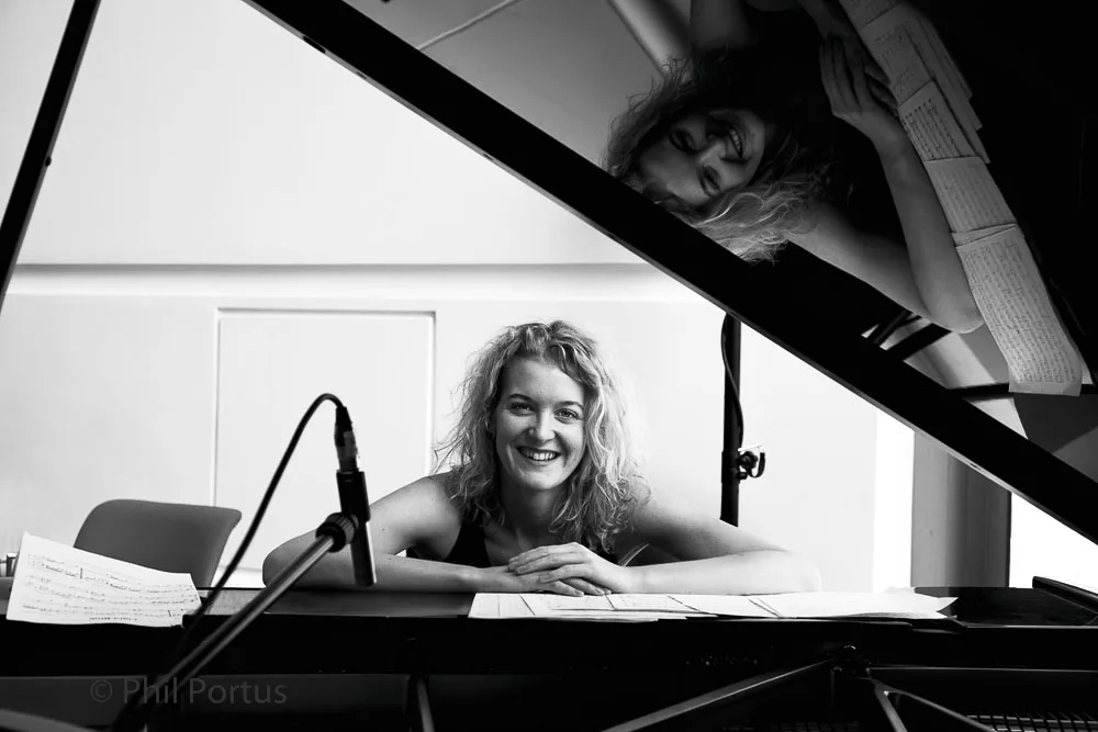 Two people are positioned near a grand piano with sheet music visible on the stand and held in one person’s hands.