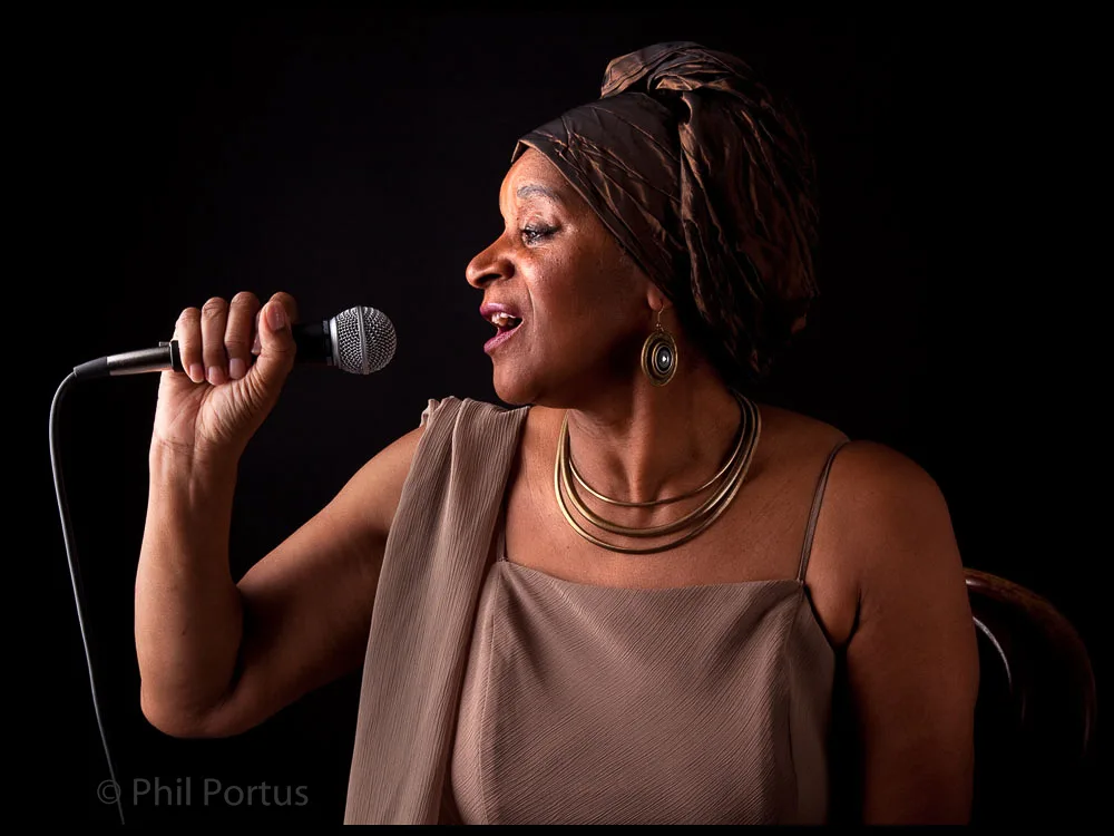 A woman sings into a microphone while wearing a headscarf and gold jewelry against a dark background.