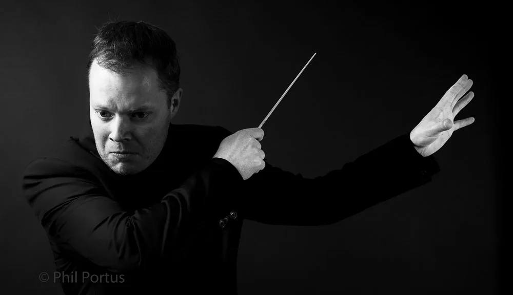 A person wearing a dark suit holds a baton while gesturing with their arm in a black and white portrait.