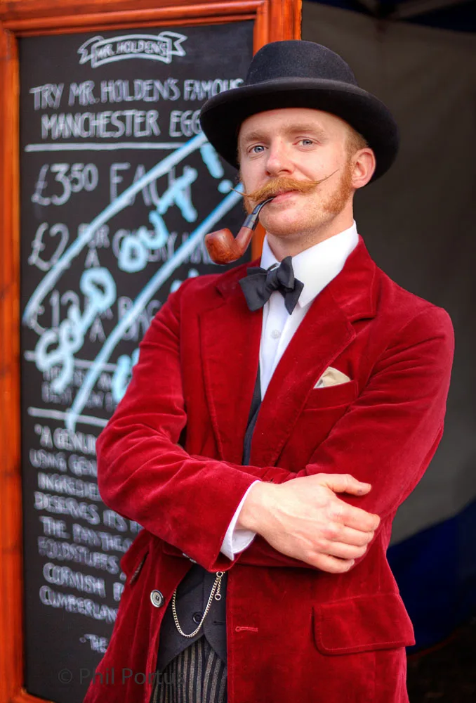 A person with a mustache and bowler hat stands with arms crossed in front of a chalkboard.
