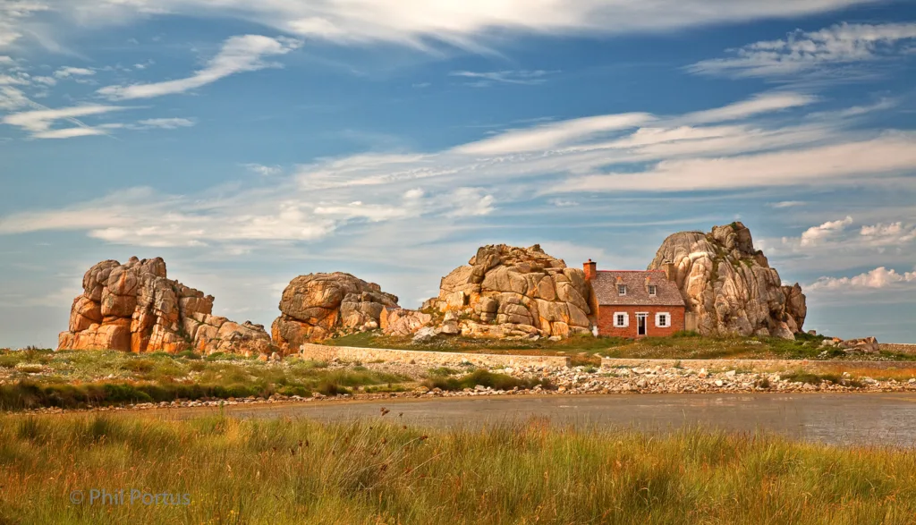 A small brick house sits nestled among large rock formations on a grassy peninsula under a partly cloudy sky.