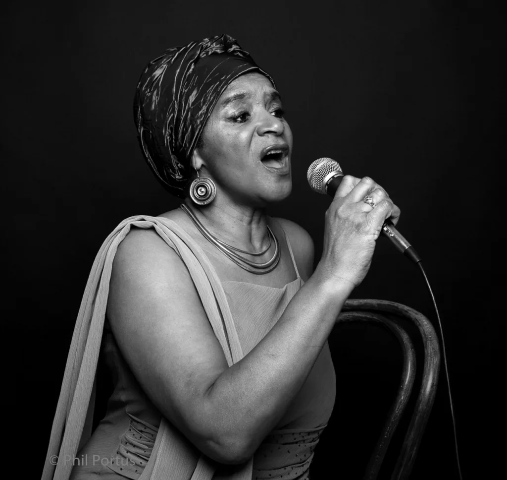 A woman sings into a microphone while seated, wearing a headscarf and jewelry.