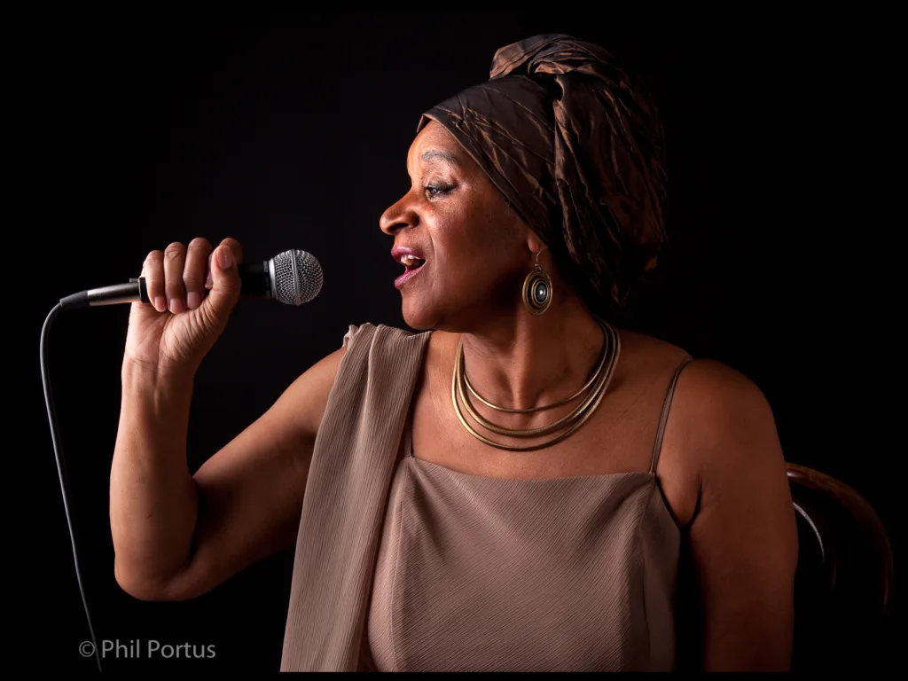 A woman sings into a microphone against a dark background, wearing a headscarf and jewelry.