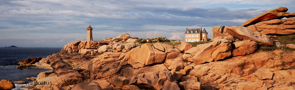 A lighthouse stands on a rocky promontory overlooking the ocean under a cloudy sky.