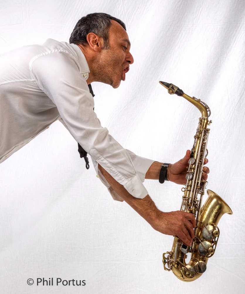 A person with graying hair plays a brass saxophone while wearing a white button-down shirt.