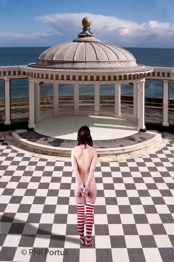 A person stands on a checkered floor facing a white domed structure overlooking water under a partly cloudy sky.