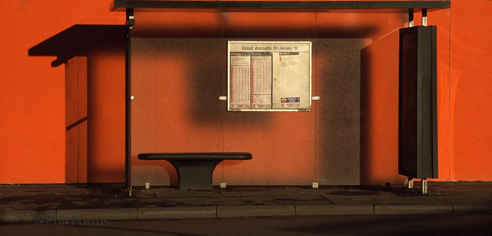 A bus shelter with a bench and timetable is illuminated by a warm light against a dark background.