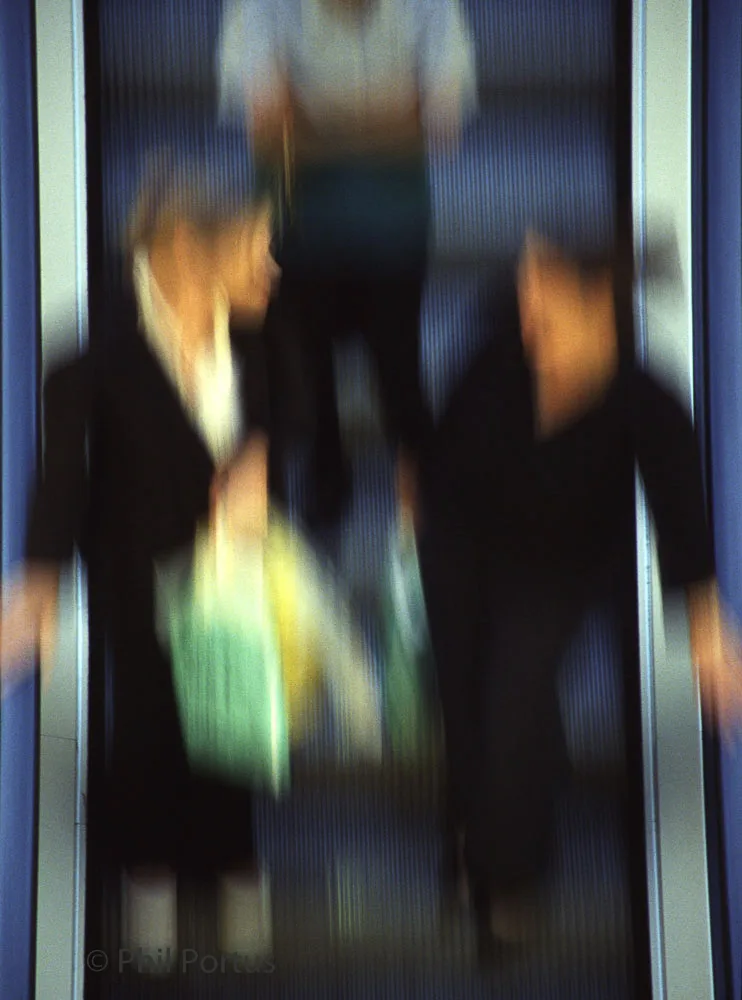 Blurred figures move upwards on an escalator within a brightly lit interior space.