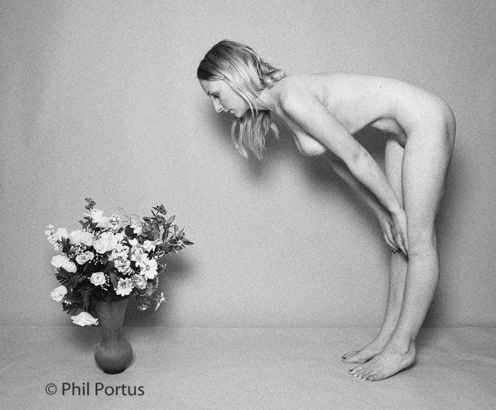 A nude person bends toward a vase of flowers in black and white.