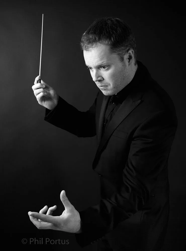 A man in a black suit holds a baton while gesturing with his hand.