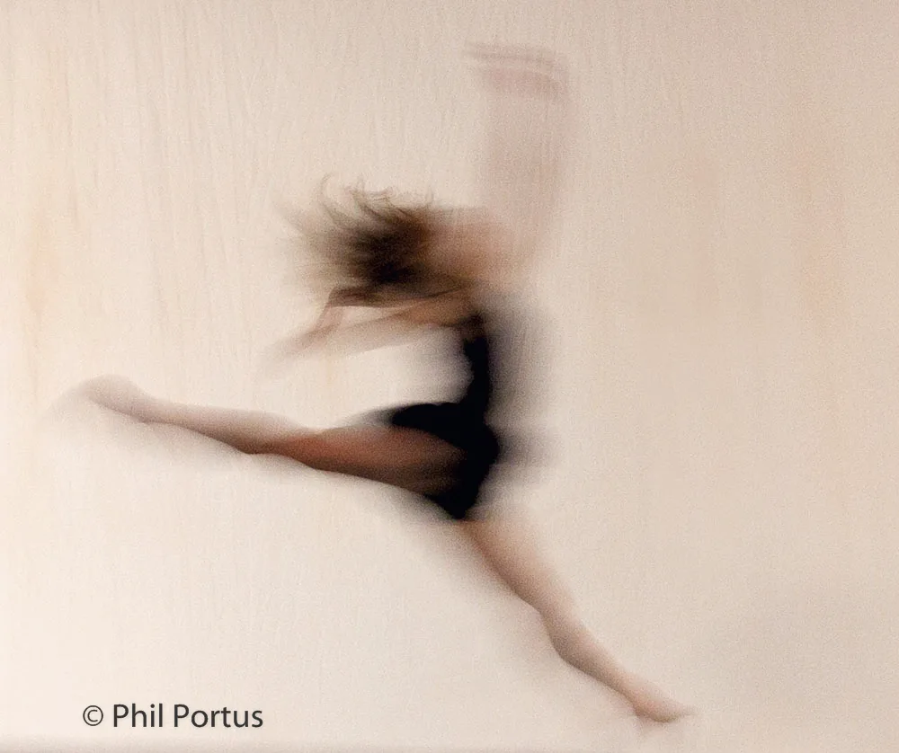 A blurred figure appears to be dancing or moving quickly against a plain light-colored background.