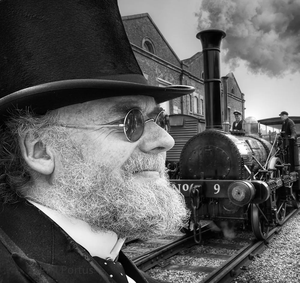 A man with a full beard and top hat looks over his shoulder at a black and white steam train.