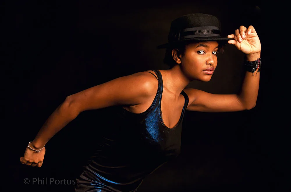 A person with dark skin adjusts a hat while striking a pose against a dark background.