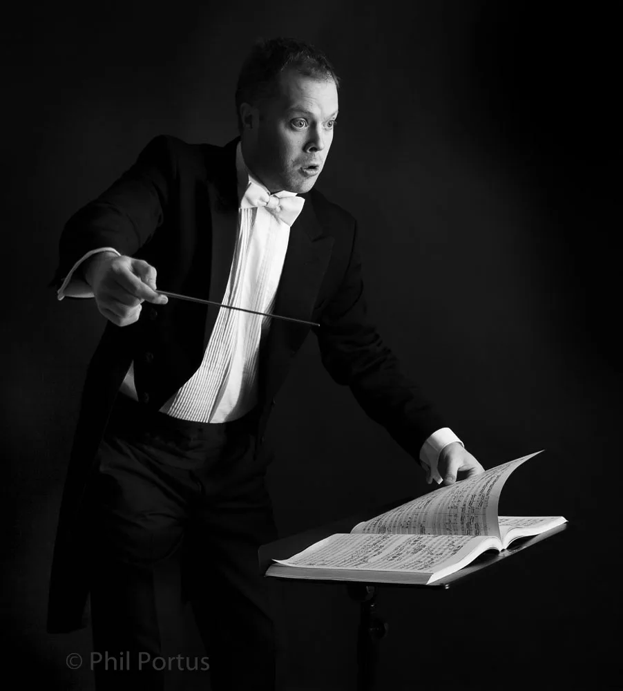 A man in formal wear conducts with a baton, standing next to an open music stand.