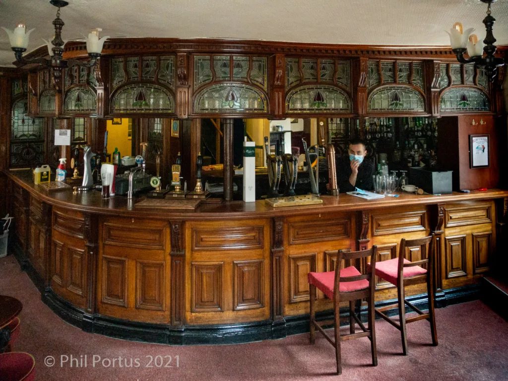 A curved wooden bar with multiple taps stands behind a person wearing a face mask.