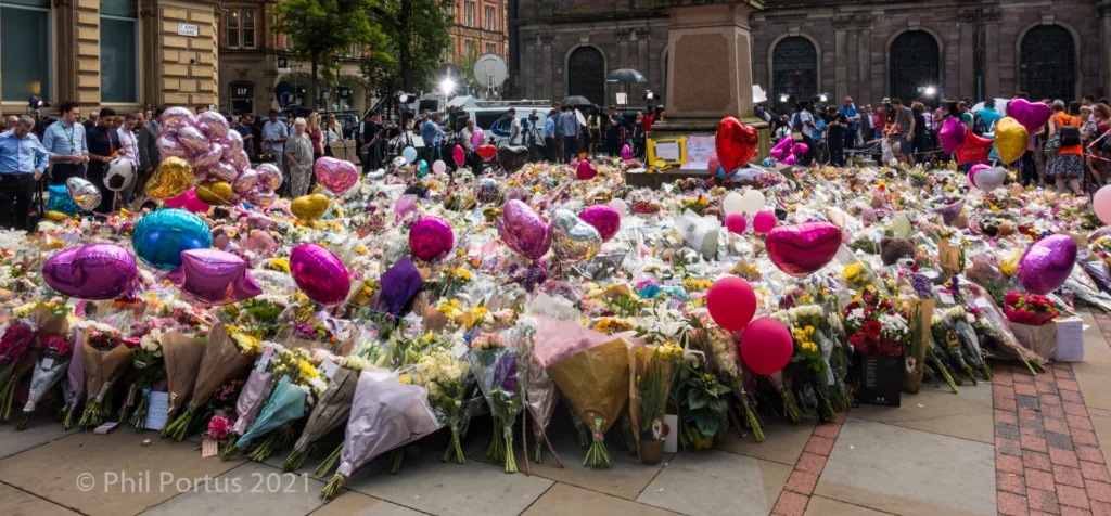 A large collection of flowers and balloons covers a paved area in front of a stone building.