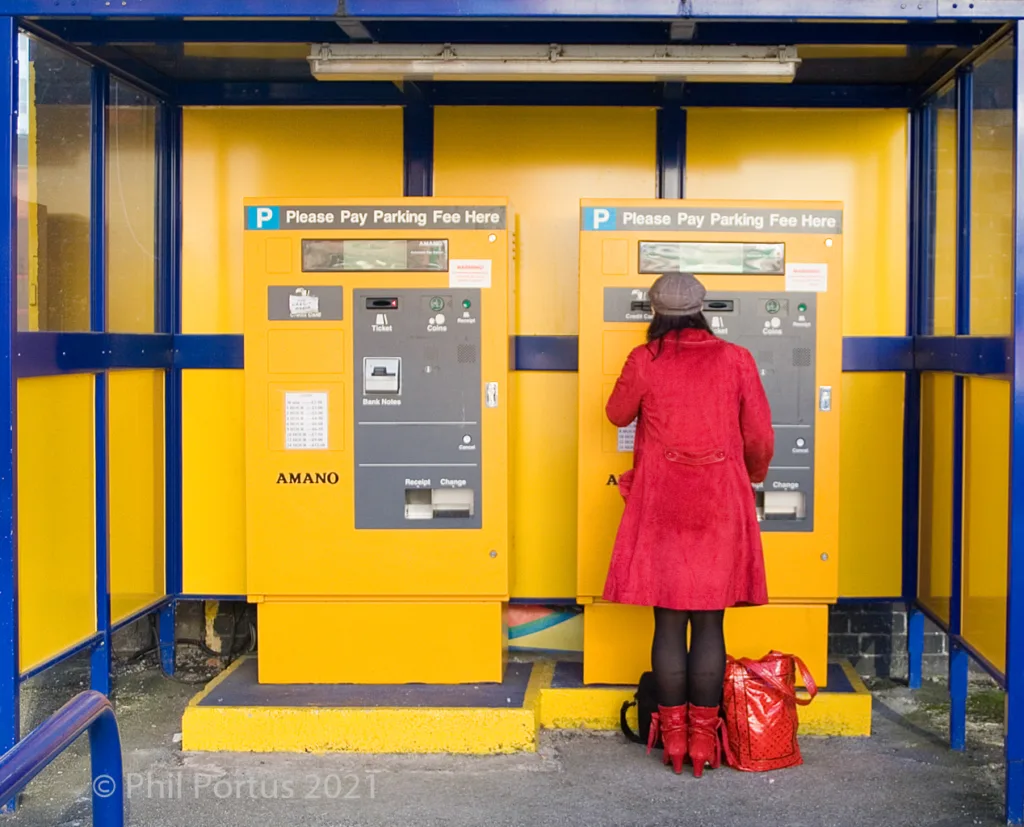 A person in a red coat uses a bright yellow parking meter enclosed by blue metal framing.