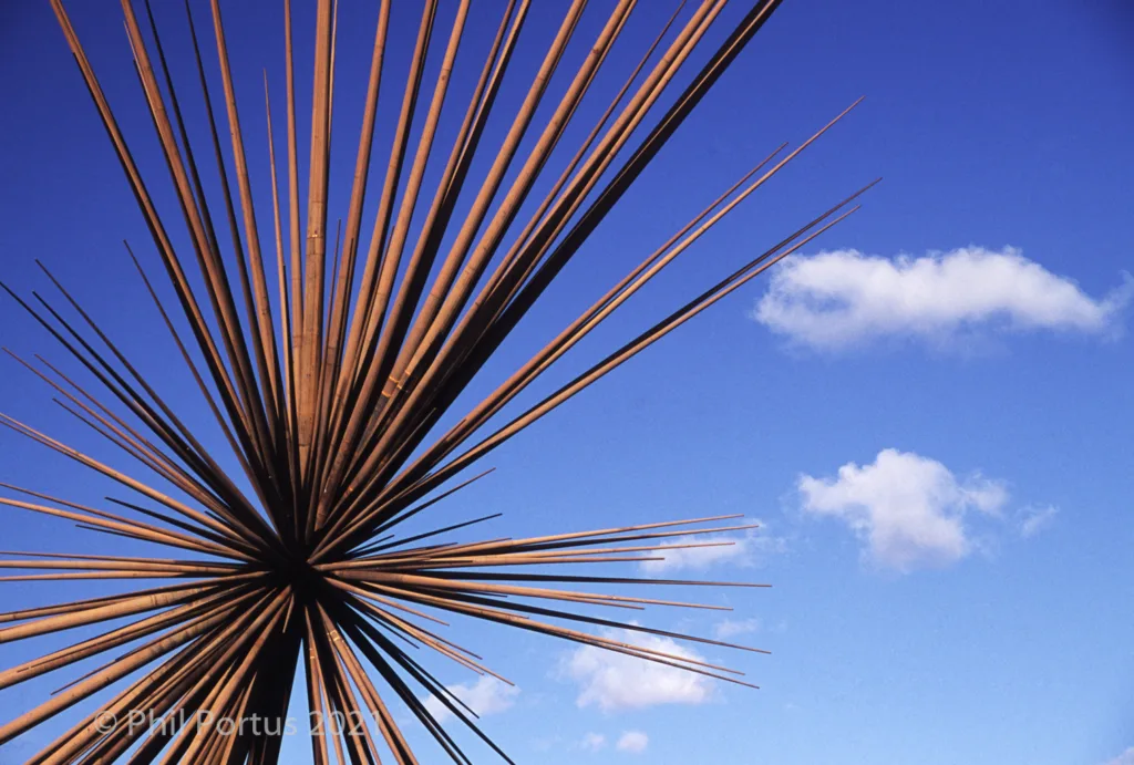 A metal sculpture with many radiating spikes extends upward against a bright blue sky with scattered clouds.