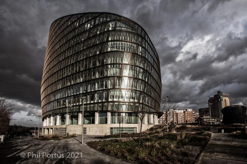 A modern curved building with glass windows stands against a dramatic sky filled with dark clouds.