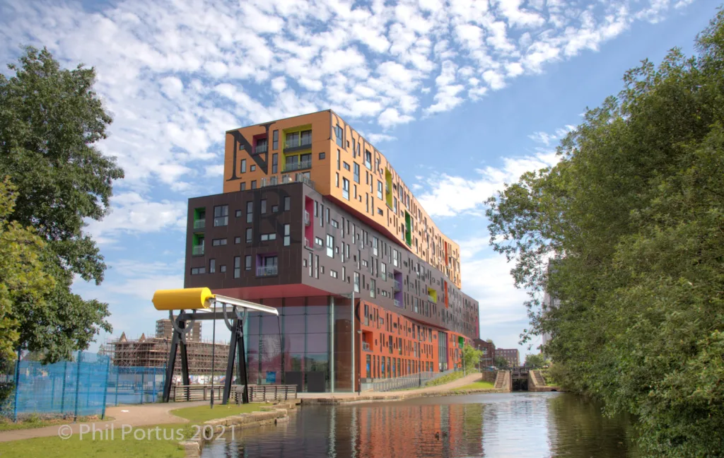 A modern building with colorful facades rises above a canal on a sunny day.