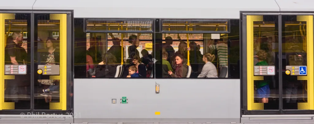 Passengers sit inside a light rail vehicle with large windows reflecting the surrounding environment.